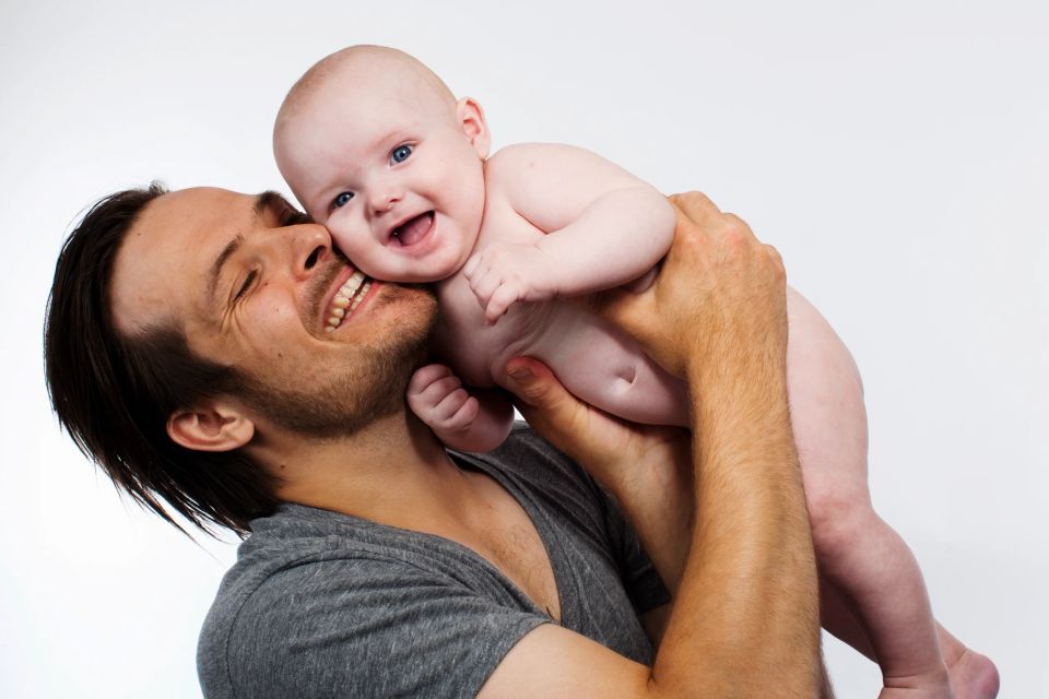 Far i grå t-shirt holder en smilende baby tæt med et stort grin på hans ansigt, i et naturligt baby foto.