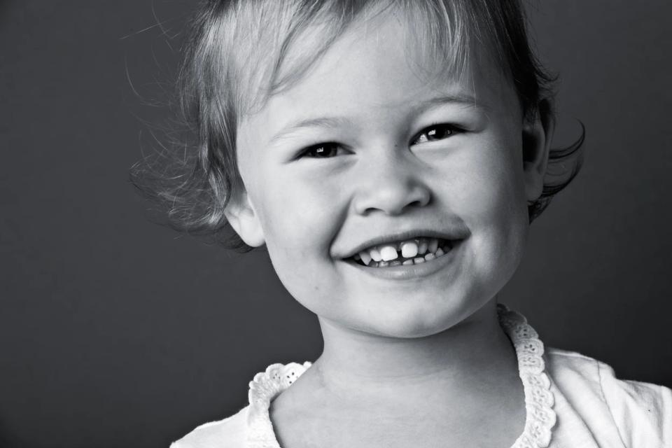 Sort/hvidt børnefotografi af en glad, smilende pige med lyst hår og kighul i tænderne, set tæt på.