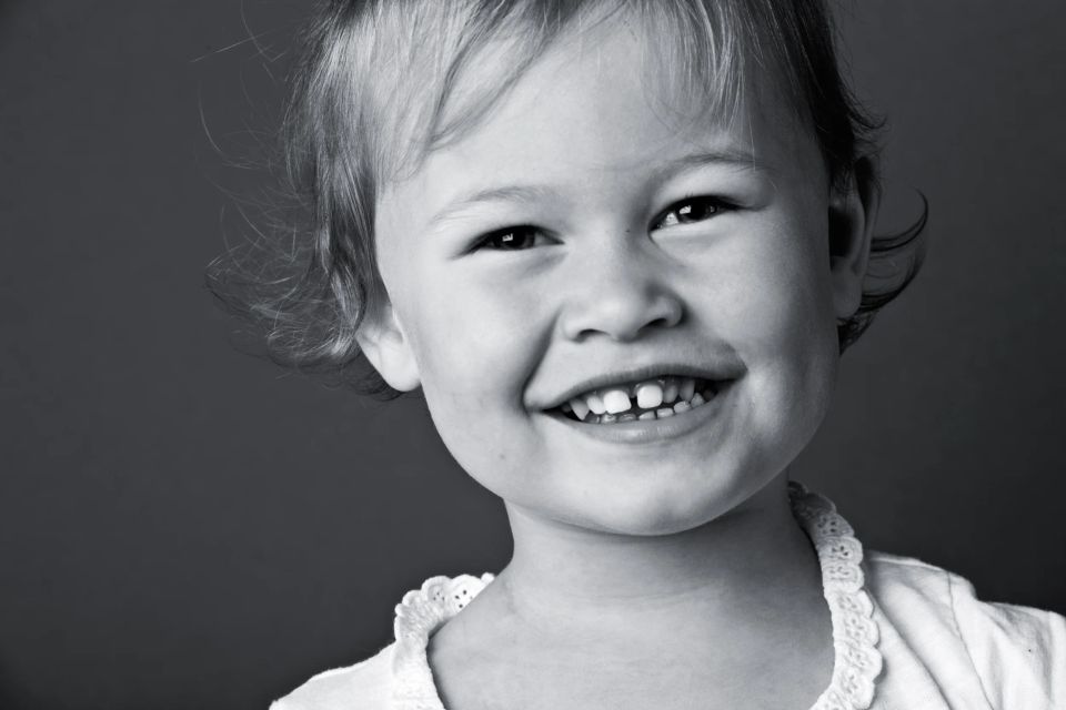 Sort/hvidt børnefotografi af en glad, smilende pige med lyst hår og kighul i tænderne, set tæt på.