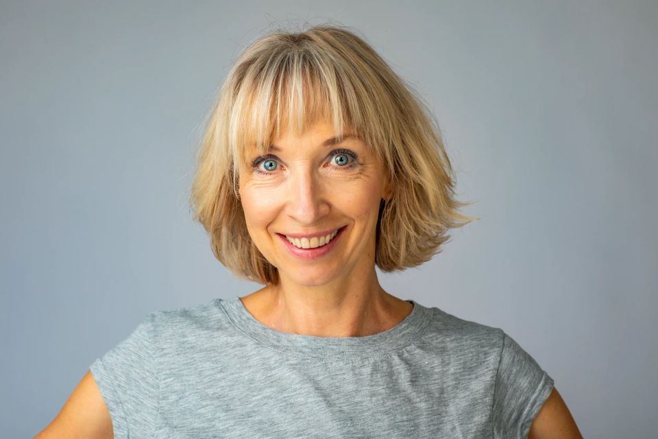 Nærbillede af smilende kvinde med kort, lyst hår og dybblå øjne i et autentisk portrætfoto med grå baggrund.