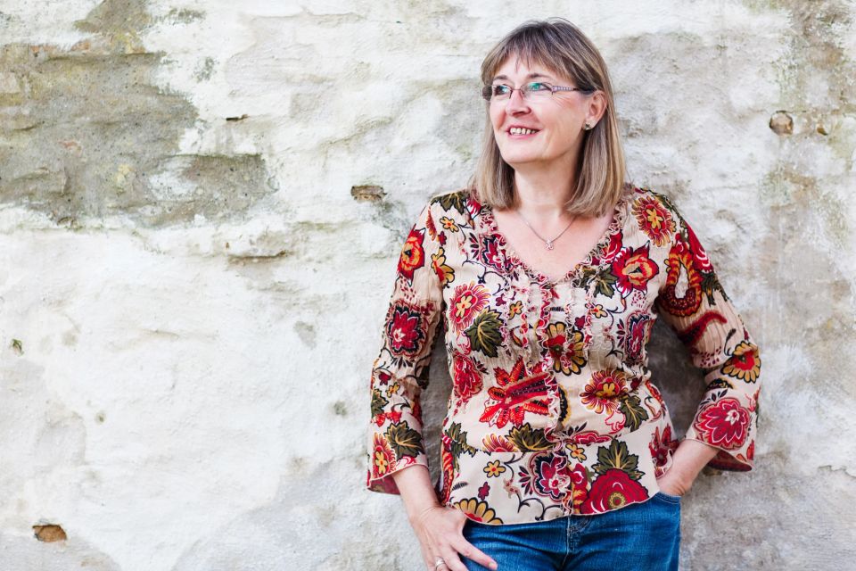 Portrætfoto af smilende kvinde med bobfrisure og blomstret bluse, der står lænet op ad en rå mur, kigger mod højre.