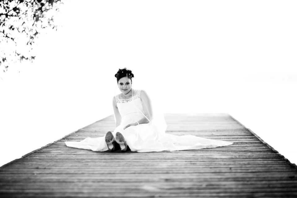 Bryllupsfoto: Smilende brud i hvid kjole sidder på en træmole i blødt dagslys med bare fødder. Naturlig stemning.