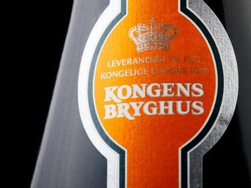 Nærbillede af orange etiket fra Kongens Bryghus med en krone og teksten "Leverandør til Det Kongelige Danske Hof" på en fl...