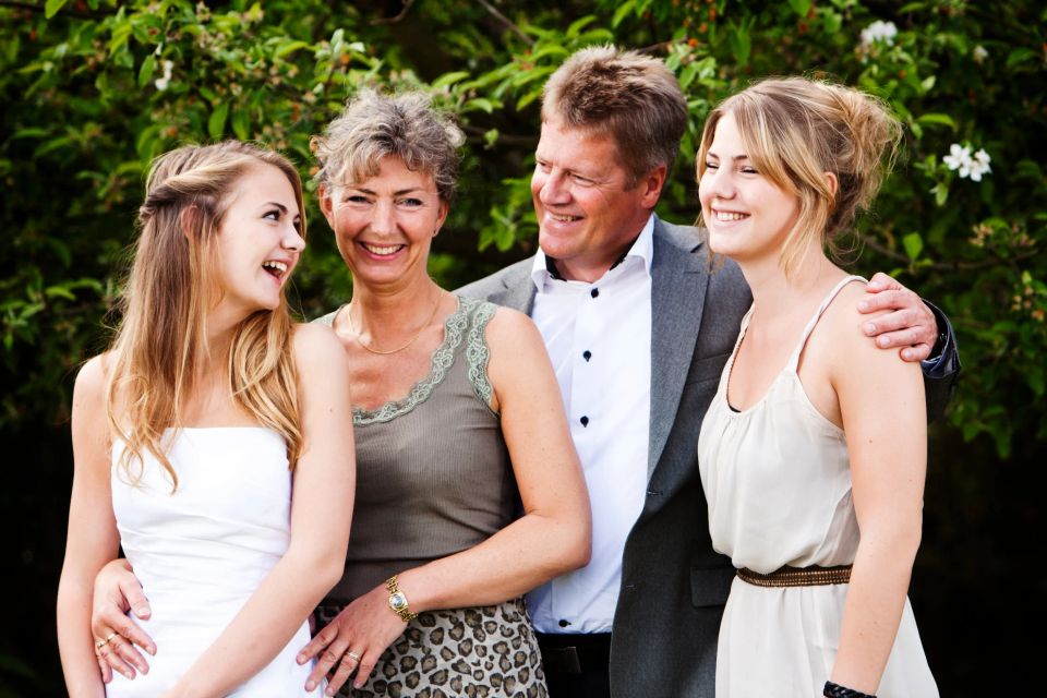 Glad familie fotograferet udendørs med grønne buske i baggrunden. Konfirmanden i hvid kjole smiler mod sin mor.