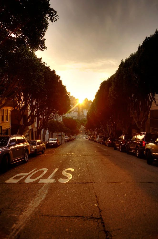 Solnedgang over en gade i San Francisco med et “STOP” skilt på vejen og parkerede biler langs vejkanten. Andet foto.