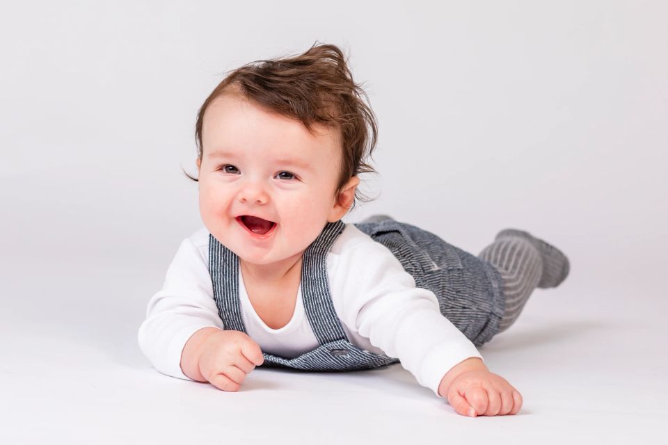 Glad baby i stribede seler og hvid trøje kravler mod beskuer i naturligt lys. Babyfotografering med fokus på glæde.