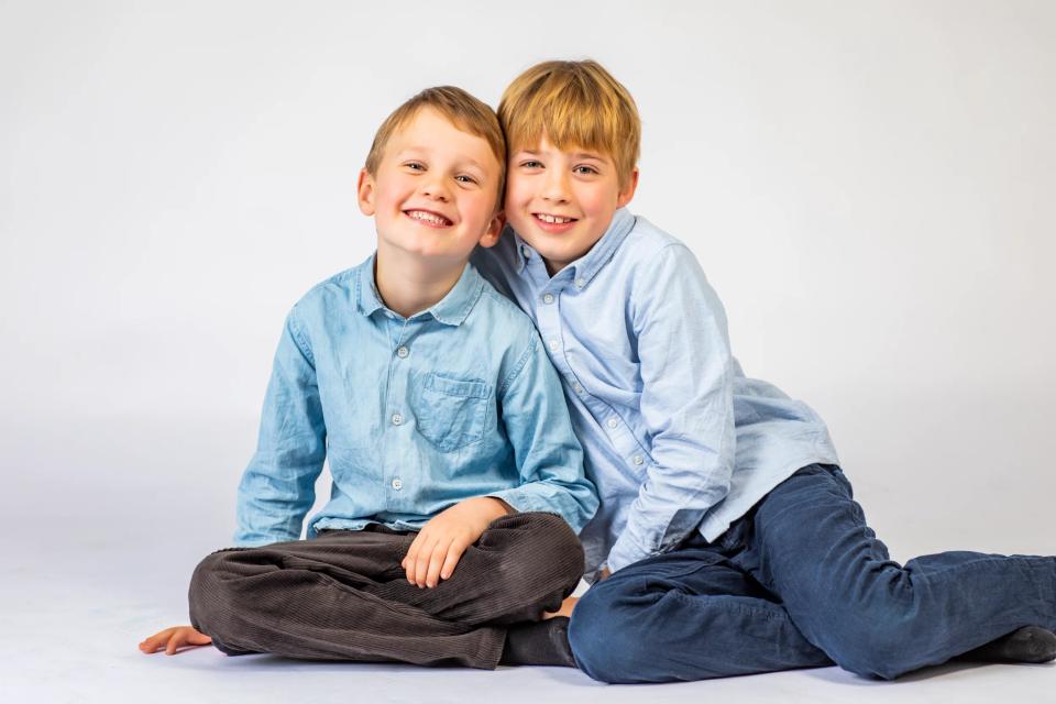 To glade drenge med lyst hår og smilende ansigter i børnefotografering, siddende tæt foran hvid baggrund.