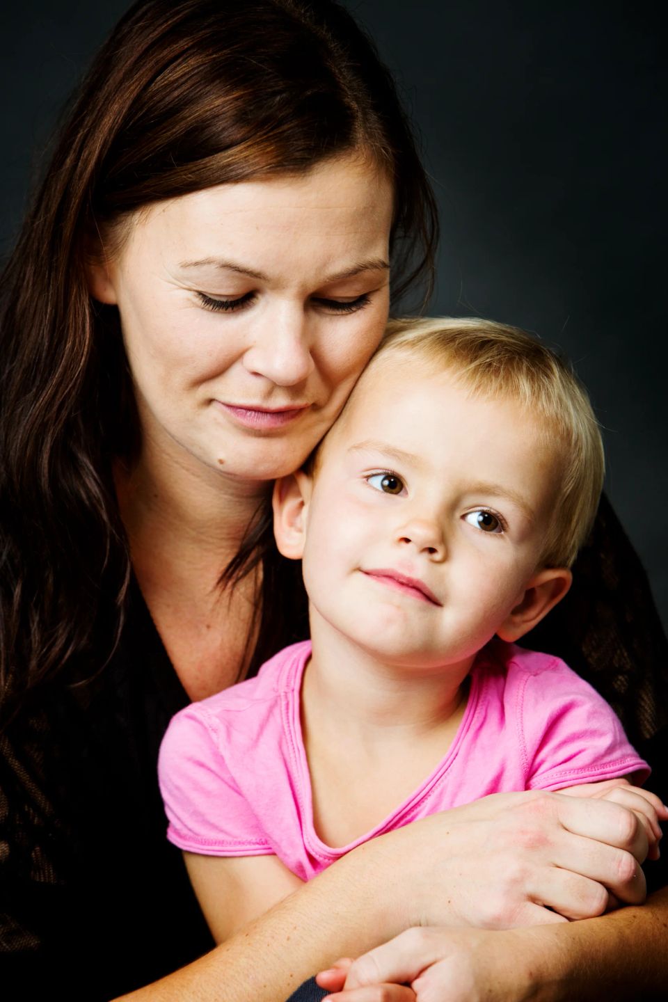 Familiefoto af mor med langt brunt hår og barn med lyst kort hår i lyserød bluse. De kigger trygt ind i kameraet.