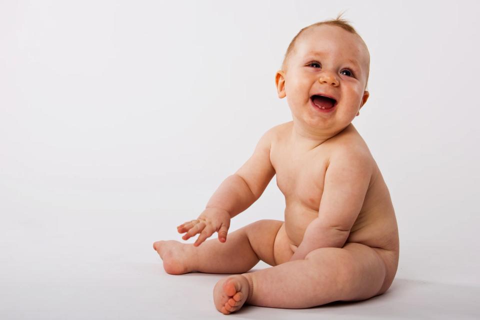 Glad babyfotografering af en grinende baby uden tøj på, siddende i klart, naturligt lys mod en simpel hvid baggrund.