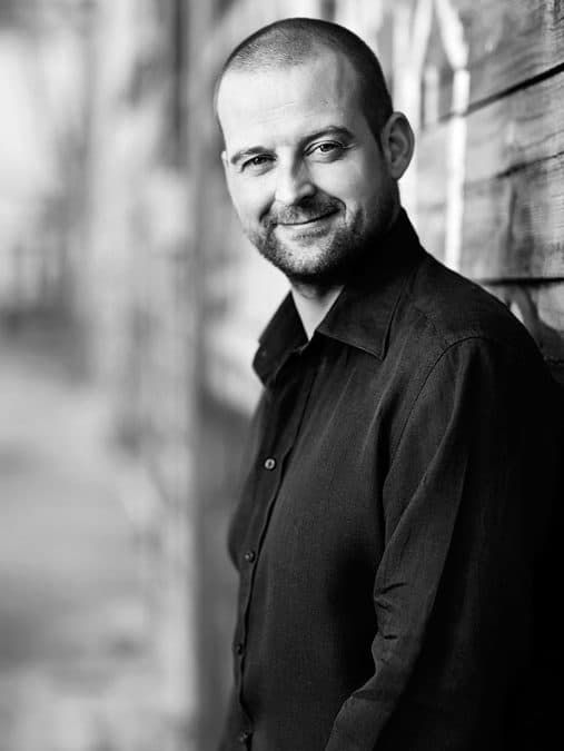 Sort/hvid portrætfoto af smilende mand med lyst skæg, iført sort skjorte, lænende sig mod trævæg