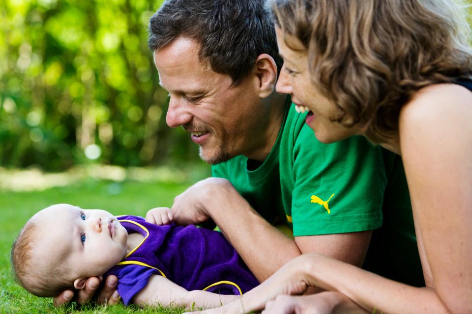 Glad babyfotografering af forældre der smilende kigger forelsket på deres baby liggende i det grønne græs.
