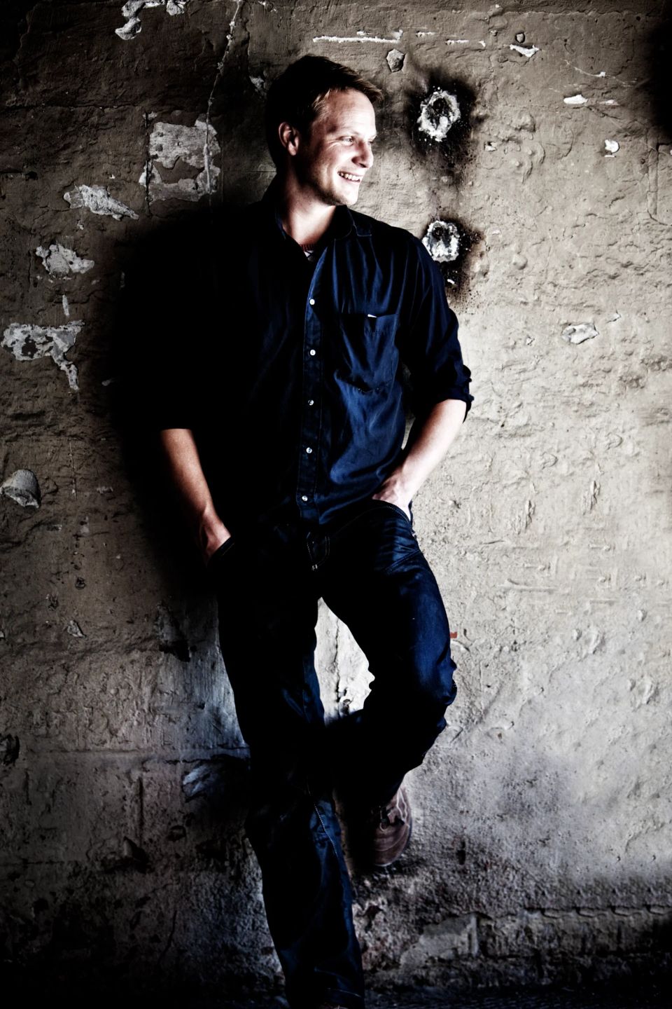 Portrætfoto af smilende mand i mørkeblå skjorte og jeans, lænet op ad en rå mur. Naturligt portræt i dagslys.