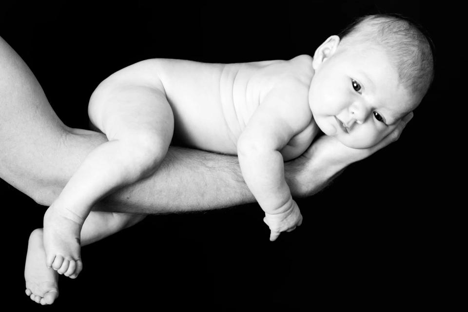 Babyfotografering i sort-hvid af nyfødt baby, der ligger trygt på en voksenarm mod sort baggrund.