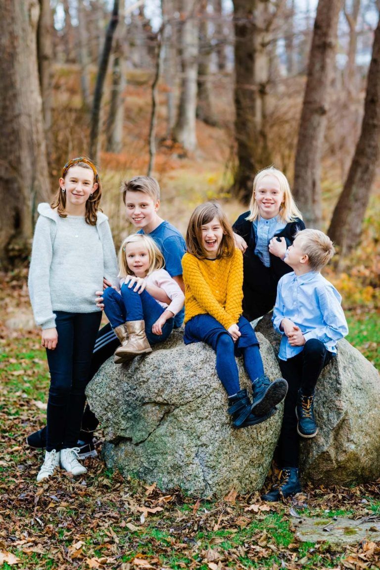 Glad familie i naturen. Seks børn sidder og står på sten i skov i varmt efterårslys, flere smiler bredt til kameraet.