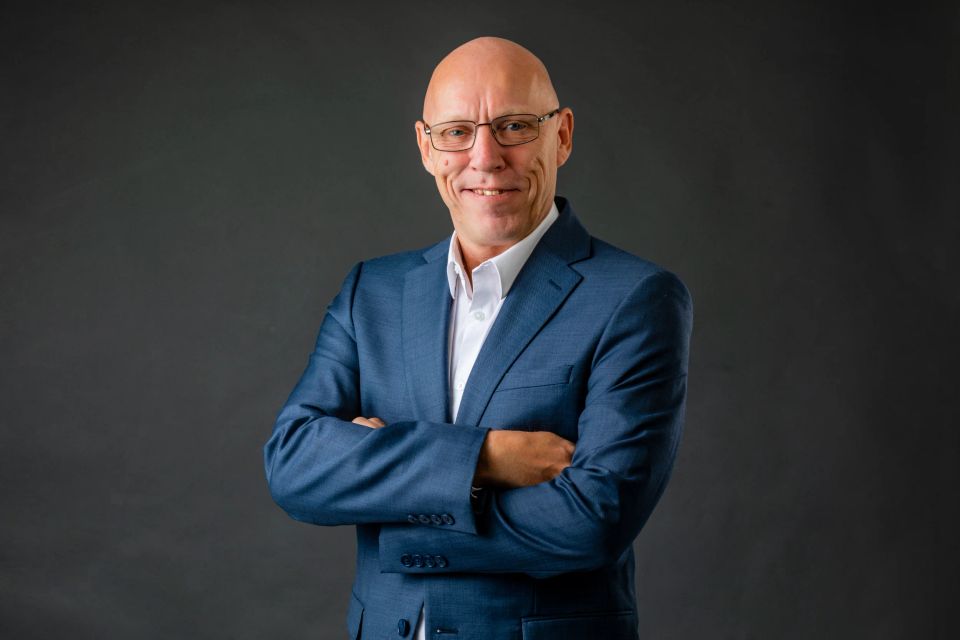 Studieportræt af smilende, skaldet mand i briller og blåt jakkesæt med armene krydset, mod en mørk baggrund.