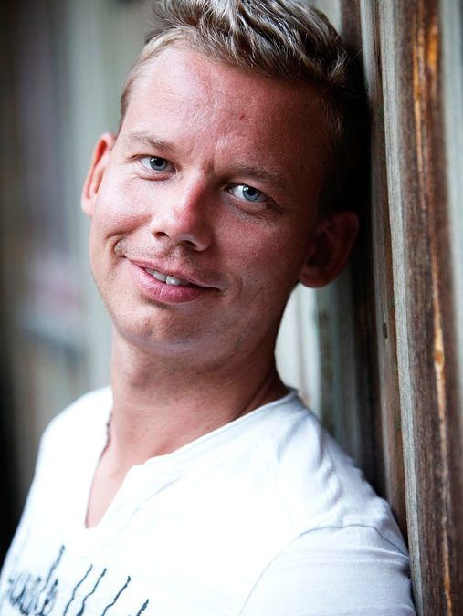 Portrætfoto af ung mand med lyst hår og blå øjne, der smiler skævt mod en trævæg.