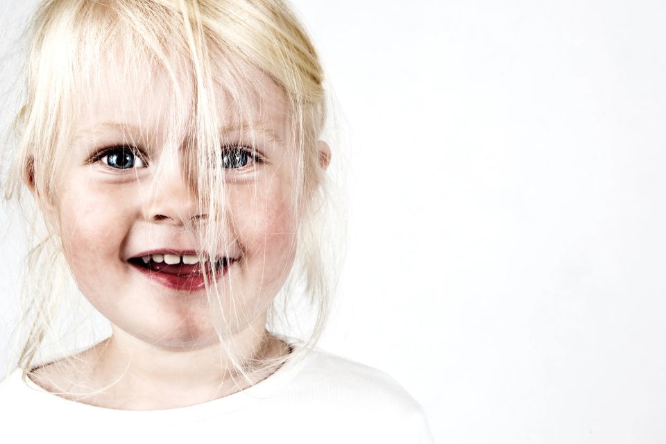 Portrætfoto af smilende pige med lyst hår, der dækker dele af ansigtet, og klare blå øjne på lys baggrund.