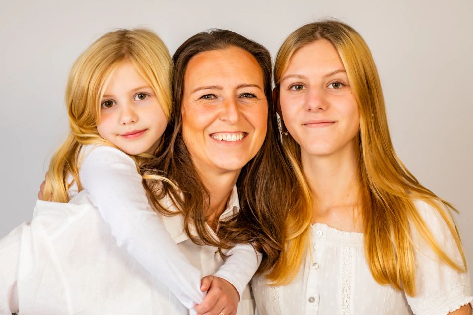 Familiefotografering: Smilende kvinde knuser glad datter, mens ældre datter står ved siden af, alle i hvide bluser.