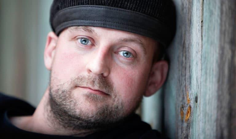Nærbillede af mand med blå øjne og hat, der læner sig mod en trævæg, i et intenst portrætfoto med blødt lys.