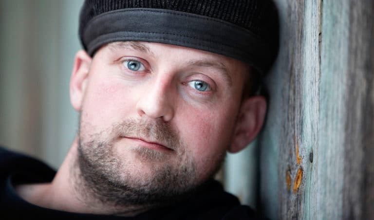 Nærbillede af mand med blå øjne og hat, der læner sig mod en trævæg, i et intenst portrætfoto med blødt lys.
