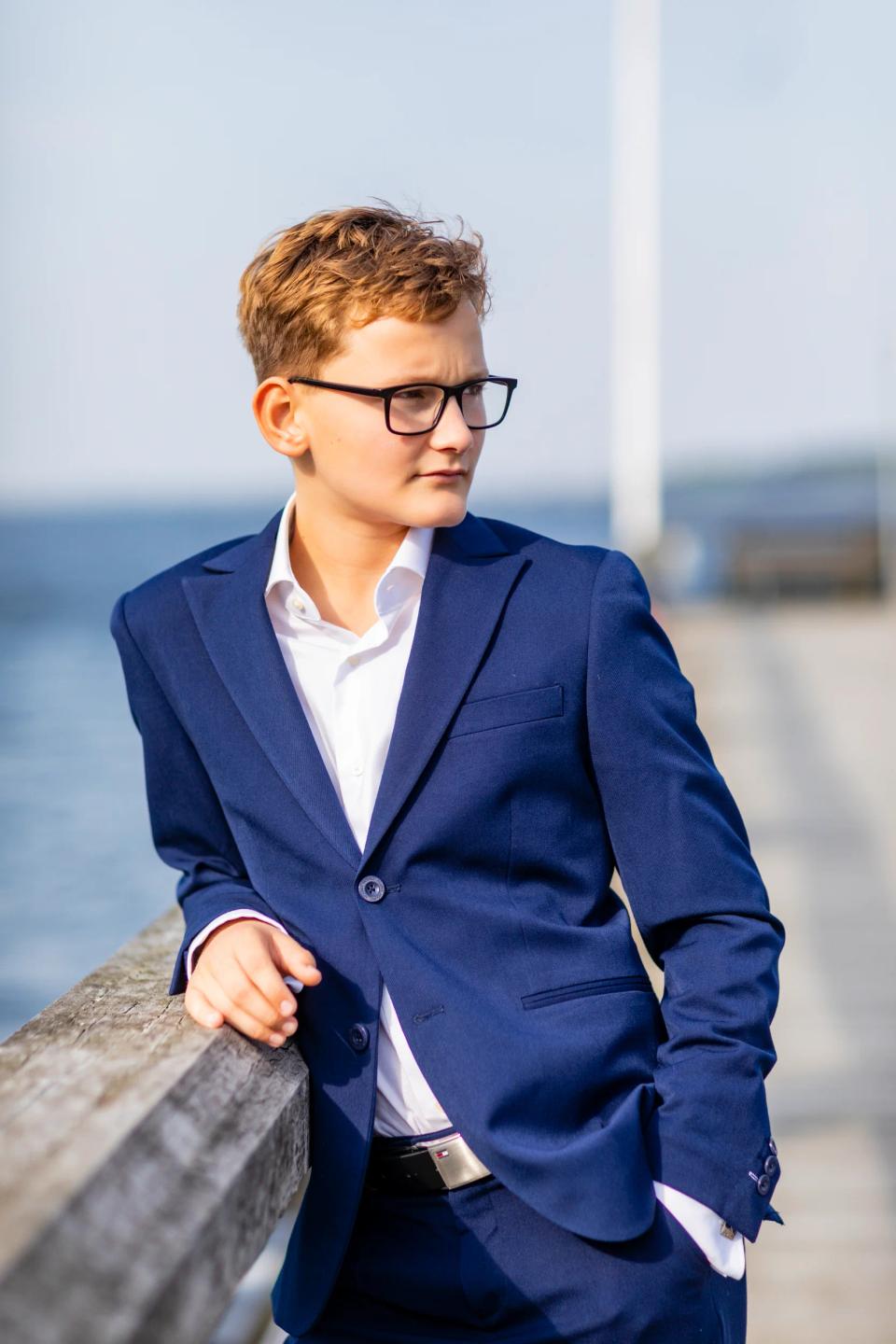 Ung konfirmand i mørkeblåt jakkesæt og hvid skjorte står ved trægelænder og kigger mod højre, med vand i baggrunden.