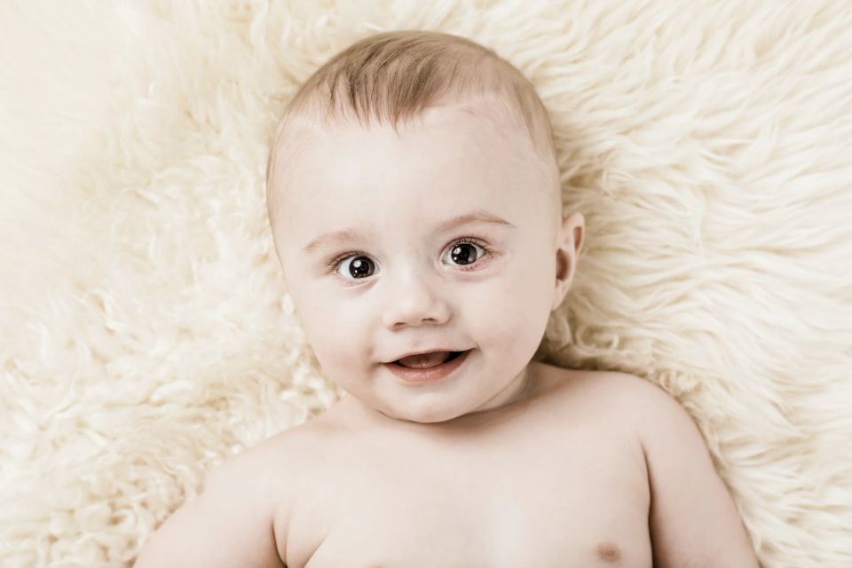 Nærbillede af glad, smilende baby med store øjne, der ligger på et lyst, blødt fåreskind. Et naturligt babyportræt.