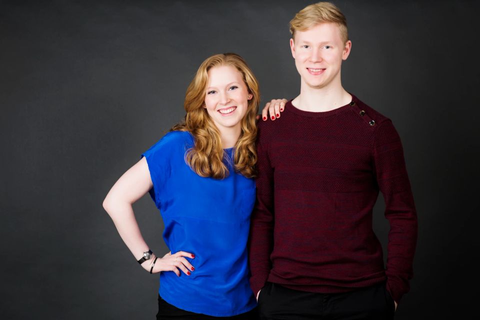 Familiefoto af smilende søskende, en kvinde i blå bluse og en mand i rød trøje, der står tæt på mørk baggrund.