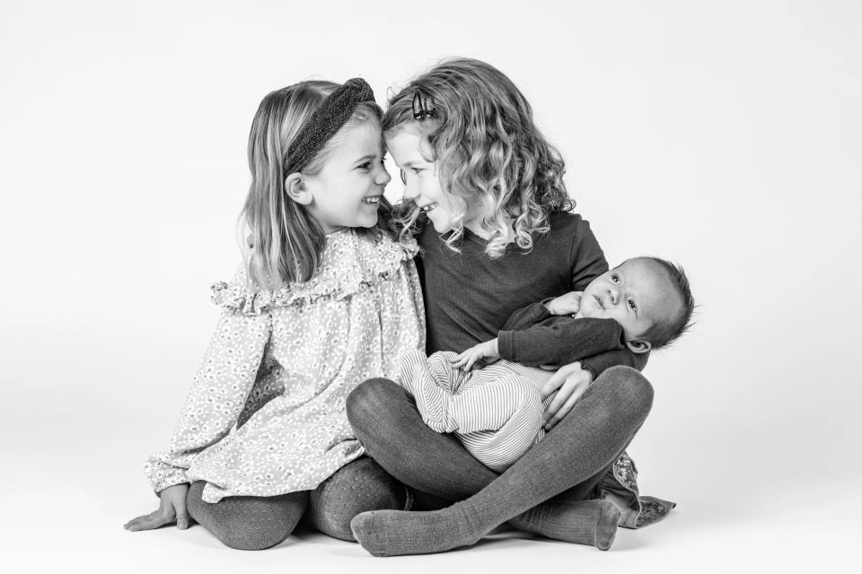 Sort-hvidt babyfoto hvor to glade søstre holder deres nyfødte lillesøster, smilende mod hinanden i blødt lys.