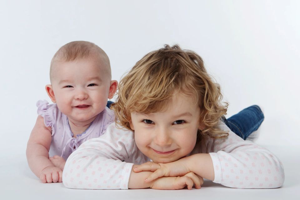 Glade søskende, en baby og et lille barn med krøller, smiler mod kameraet på et lyst studiebillede. Børn fotograferet i Vi...