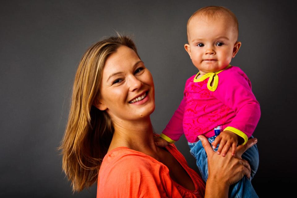 Glad mor holder smilende baby i en pink trøje. Babyfotografering med fokus på nærvær og glæde.