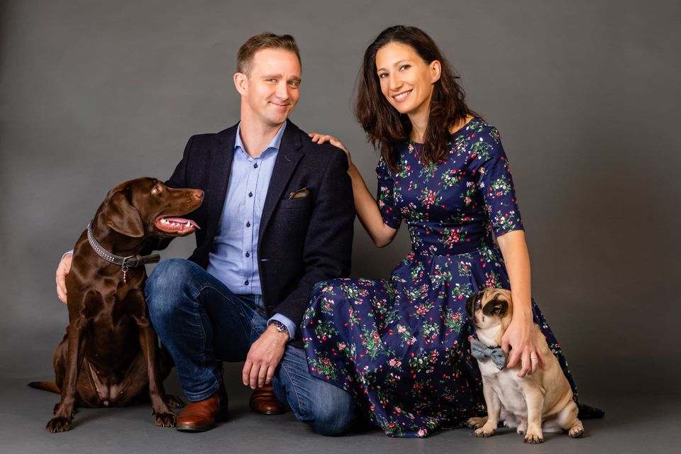 Portrætfoto af smilende par, der sidder på gulvet med deres labrador og mops foran en grå baggrund. Et ægte familiefoto.