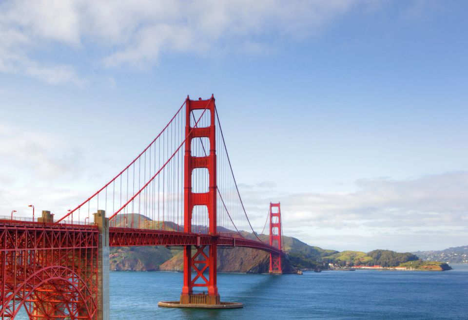 Golden Gate-broen fotograferet i oversigtsbillede over vandet med kystlinje og grønne bjerge i baggrunden under blå himmel.