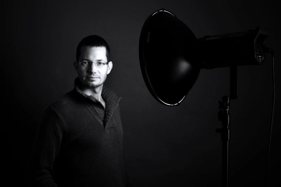 Professionelt sort-hvid portrætfoto af en mand med briller, der ser direkte ind i kameraet med et studieflash til højre.