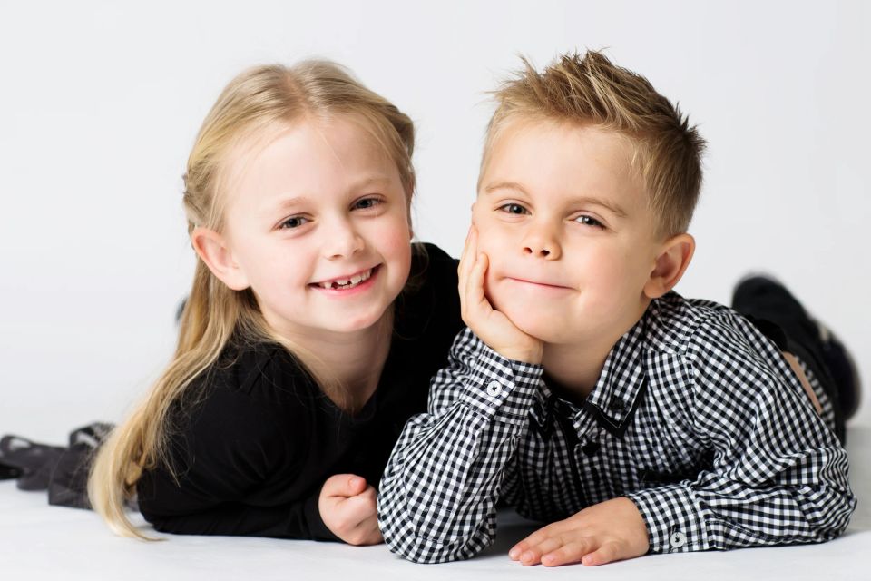 Børnefotografi af glad pige og smilende dreng liggende på maven mod hvid baggrund i blødt lys.