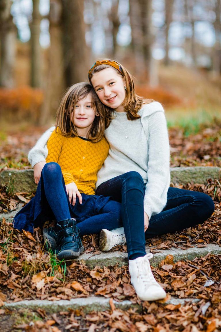 To glade børn, piger, sidder tæt sammen i efterårsblade. Et smukt fotominde fra natur nær Virum.