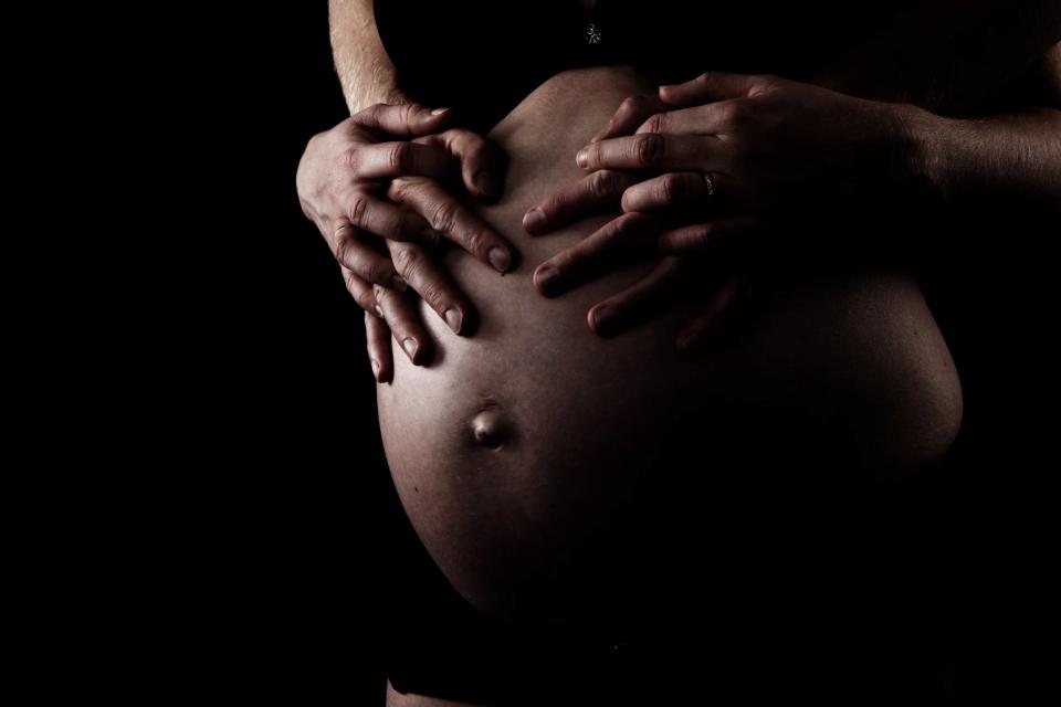 Nærbillede af gravid mave i blødt sidelys med to sæt hænder, der holder om den, symboliserende kærlighed og forventning.