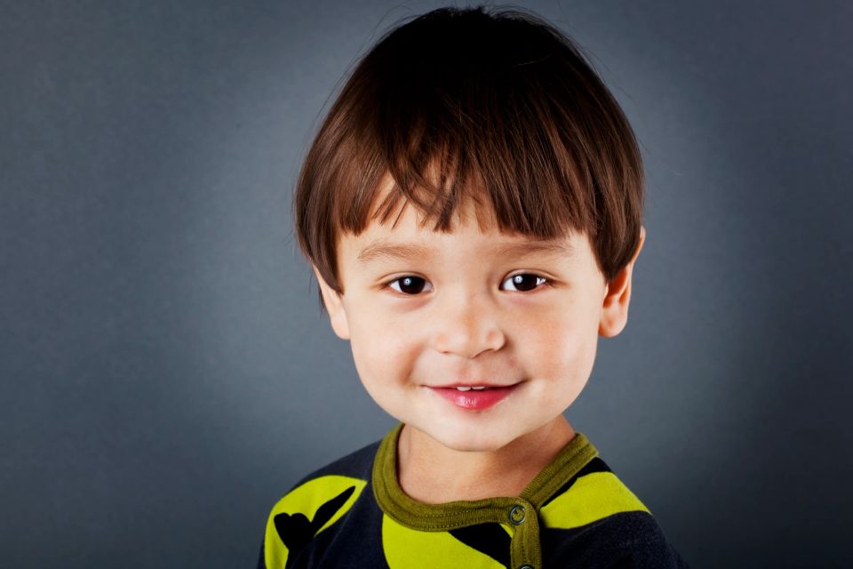 Nærbillede af en glad dreng med autentisk smil, mørkt hår og store øjne, i et børneportræt med blød belysning.