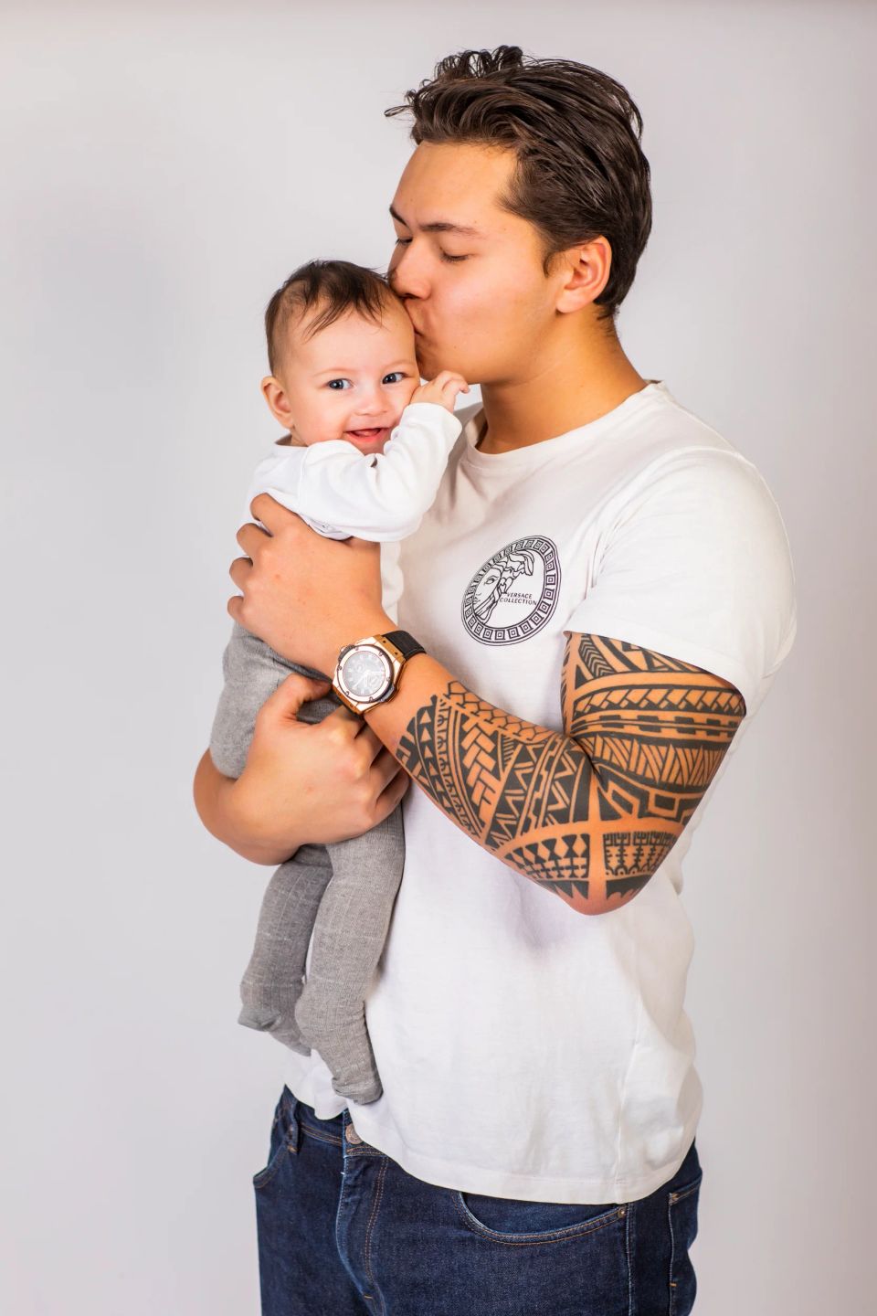 Far kysser glad baby på panden i et babyfoto. Fars arm har store tatoveringer. Kærligt øjeblik.