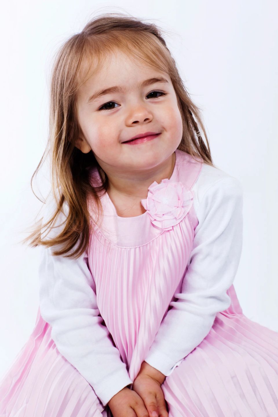 Glad lille pige i lyserød plisseret kjole med blomst på, smilende mod kameraet i et lyst børnefoto.