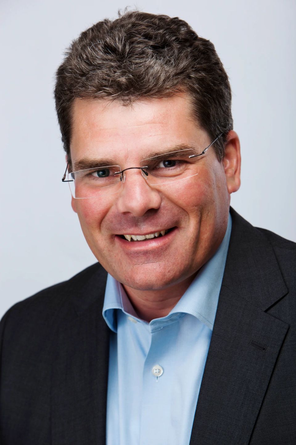 Nærbillede af smilende mand med briller i professionelt erhvervsportræt, iført mørk jakke og lyseblå skjorte.