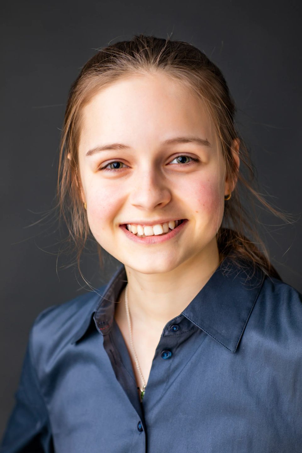 Portrætfoto af ung, smilende kvinde med lyst hår, der bærer en mørkeblå skjorte, mod en ensfarvet grå baggrund.