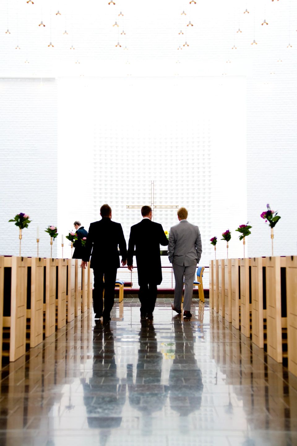 Bryllupsfotografering: Tre mænd i jakkesæt går ned ad kirkegulvet mod et stort kors i et lyst kirkerum.