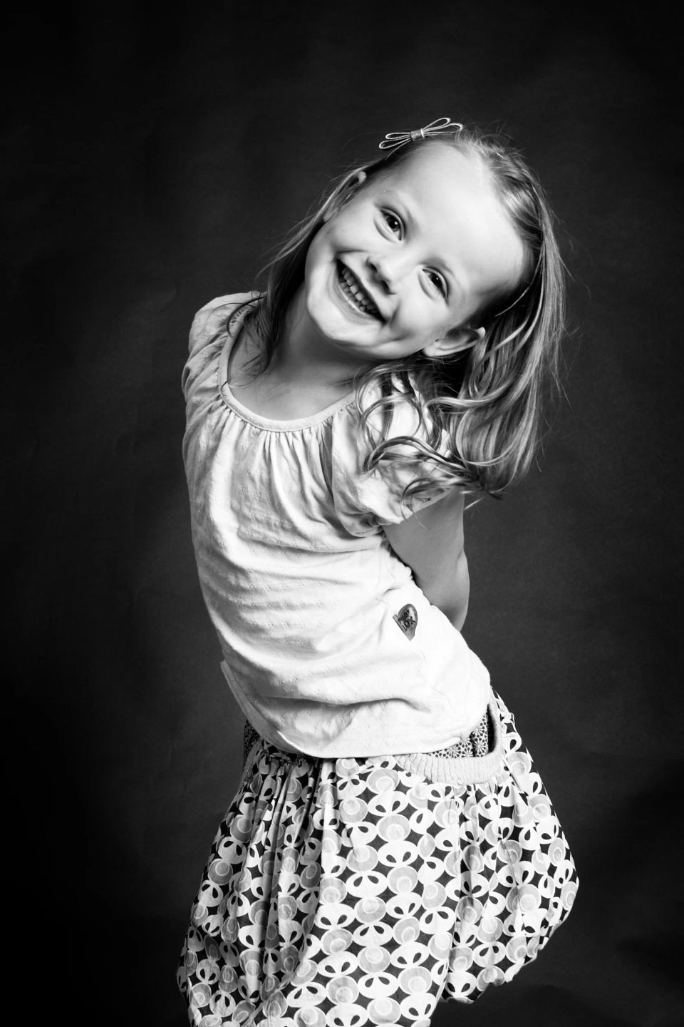 Glad, legende pige i sort-hvid fotografering, der smiler bredt med tænder i blødt lys, let bøjet i overkroppen.