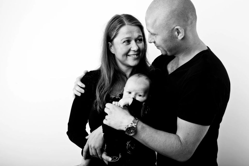 Nærportræt i sort-hvid af familie. Mor smiler til far, der holder deres spædbarn, som kigger nysgerrigt frem.