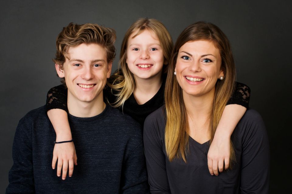 Familiefoto af tre smilende søskende med lyst hår, omfavnende hinanden foran en grå baggrund.