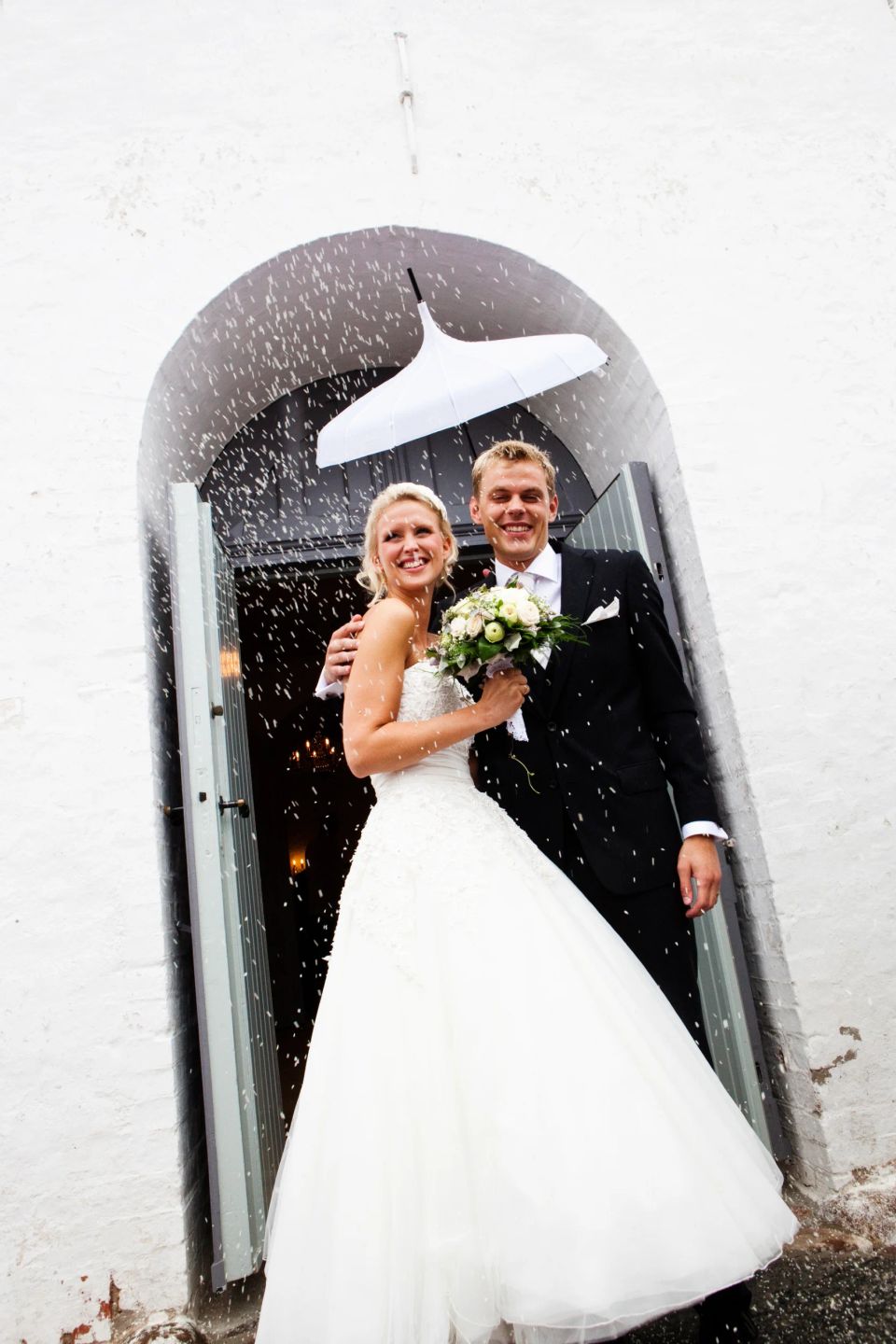 Bryllupsfoto af smilende brudepar i kirkedør med riskast. Bruden i hvid kjole, gommen i sort jakkesæt.