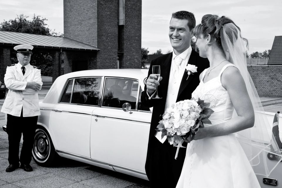 Bryllupsfotografering af smilende brudepar foran hvid veteranbil med chauffør i sort/hvidt billede.