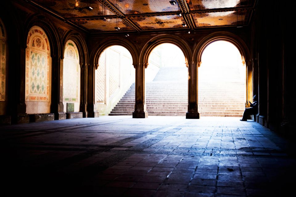 Buer og hvælvinger i en hal med en trappe i baggrunden, hvor en person sidder i varmt naturligt sidelys. Andet fotobillede.