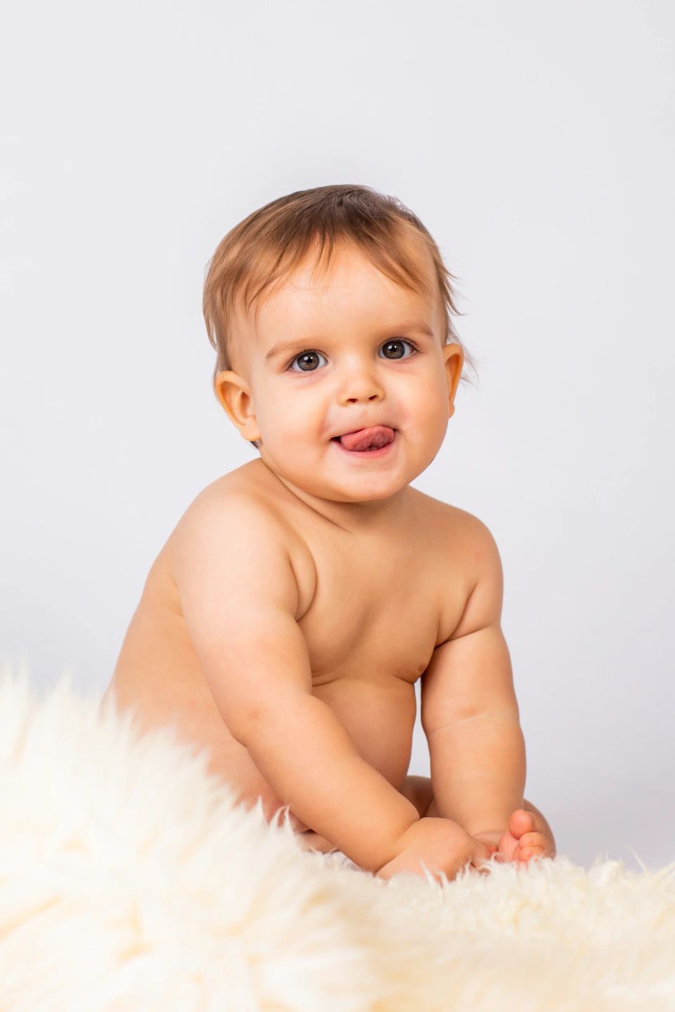 Legesyg baby med lysebrunt hår, der kigger direkte ind i kameraet med tungen ud af munden på et blødt, hvidt underlag.