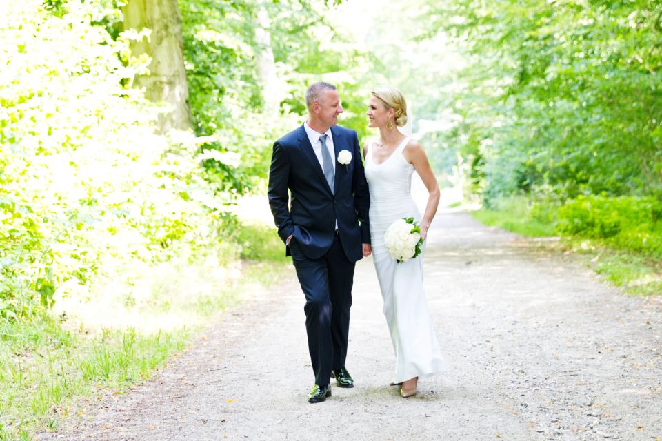 Bryllupsfotografering af smilende brudepar der går hånd i hånd på grusvej i solbeskinnet grøn skov.